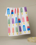 The Poppy Popsicles tea towel by Geometry features playful popsicle illustrations on white and hangs above a stack of yellow and white striped plates—perfect for adding a cheerful touch to your kitchen.