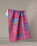The Positive Vibration tea towel by Sareka Unique, featuring a bold geometric pattern in pink, blue, orange, and red, hangs on a clothesline against a neutral background.