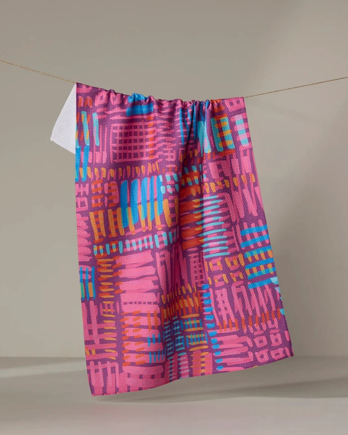 The Positive Vibration tea towel by Sareka Unique, featuring a bold geometric pattern in pink, blue, orange, and red, hangs on a clothesline against a neutral background.