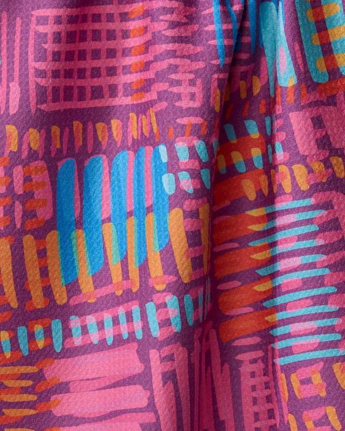 A close-up of Sareka Unique's “Positive Vibration” tea towel shows its abstract pink, orange, yellow, and blue design with textured shapes and lines on slightly folded fabric.