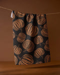 The Geometry Pumpkin Noir recycled kitchen towel, featuring an orange pumpkin pattern on black, hangs from a string against a brown background.