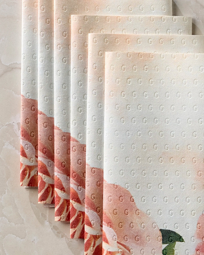 Five neatly stacked Rhapsody in Bloom reusable towels by Stefanie Bales, featuring textured sheets with a subtle G pattern and peach-pink floral blooms along the edges, displayed on a light marble surface.