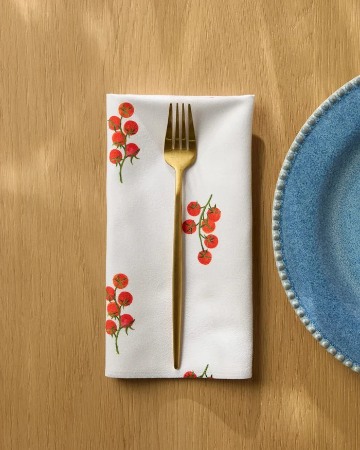 A gold fork sits on a white Geometry "Ripe and Ready Scattered" reusable dinner napkin with red tomato patterns, beside a blue ceramic plate on a light wooden table.