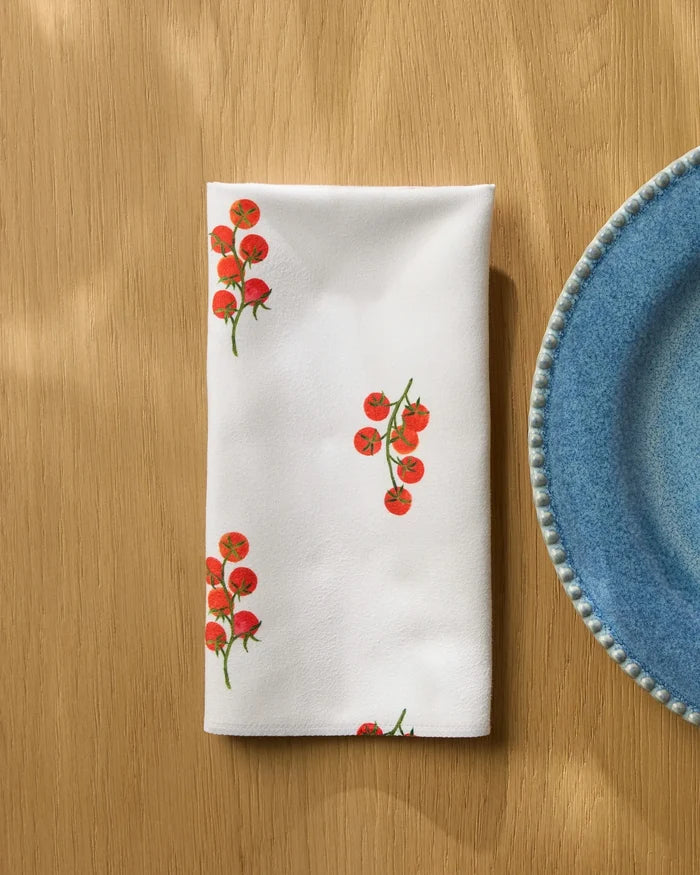 The Geometry "Ripe and Ready Scattered" white reusable napkin featuring a cherry tomato print is neatly folded on a light wooden table beside a blue ceramic plate with a textured rim.