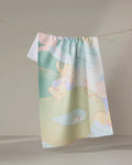 The Secret Fort dish towel by Kristina Baker, featuring pastel hues and abstract, playful animal and shape designs, hangs from a clothesline against a neutral background.