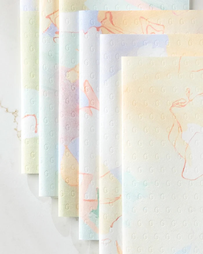 Secret Fort kitchen towels by Kristina Baker are eco-friendly and pastel-colored, with a subtle embossed “G” pattern and soft abstract swirls in light orange, blue, and green, neatly stacked on a marble surface.