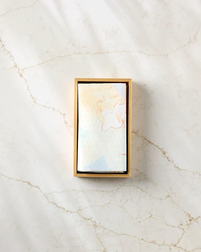 A rectangular box of Secret Fort by Kristina Baker, holding a folded, patterned napkin that resembles eco-friendly kitchen towels, rests on white marble with light beige veining.