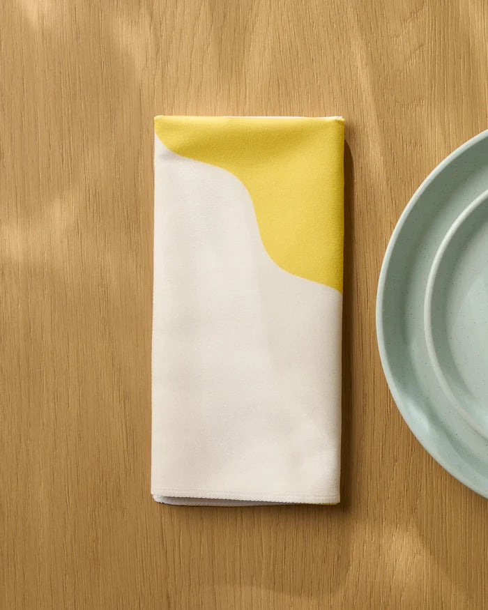 A folded Geometry Solar Light reusable napkin with a yellow and white abstract design sits on a light wood table next to two stacked pale blue plates.