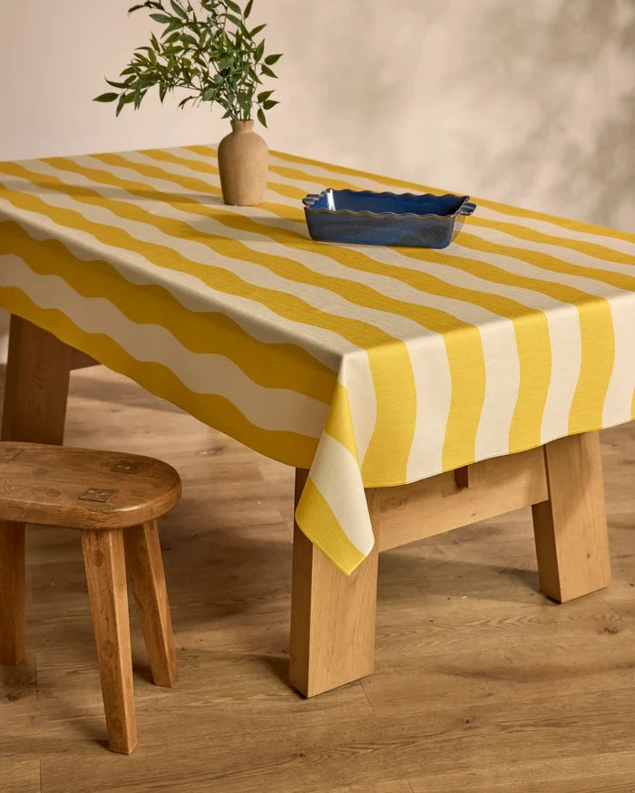 A Geometry Solar yellow and white striped tablecloth covers a wooden table set with a blue rectangular dish and a vase of green branches, placed on a wooden floor beside a rustic wooden stool.