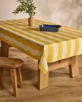 A Geometry Solar yellow and white striped tablecloth covers a wooden table set with a blue rectangular dish and a vase of green branches, placed on a wooden floor beside a rustic wooden stool.