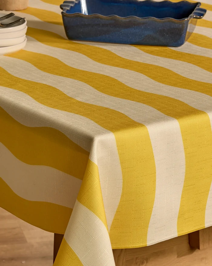 A close-up of a table with the Solar spill-resistant tablecloth by Geometry, featuring yellow and white wavy stripes. Stacked white plates and a blue rectangular ceramic dish are arranged on top.
