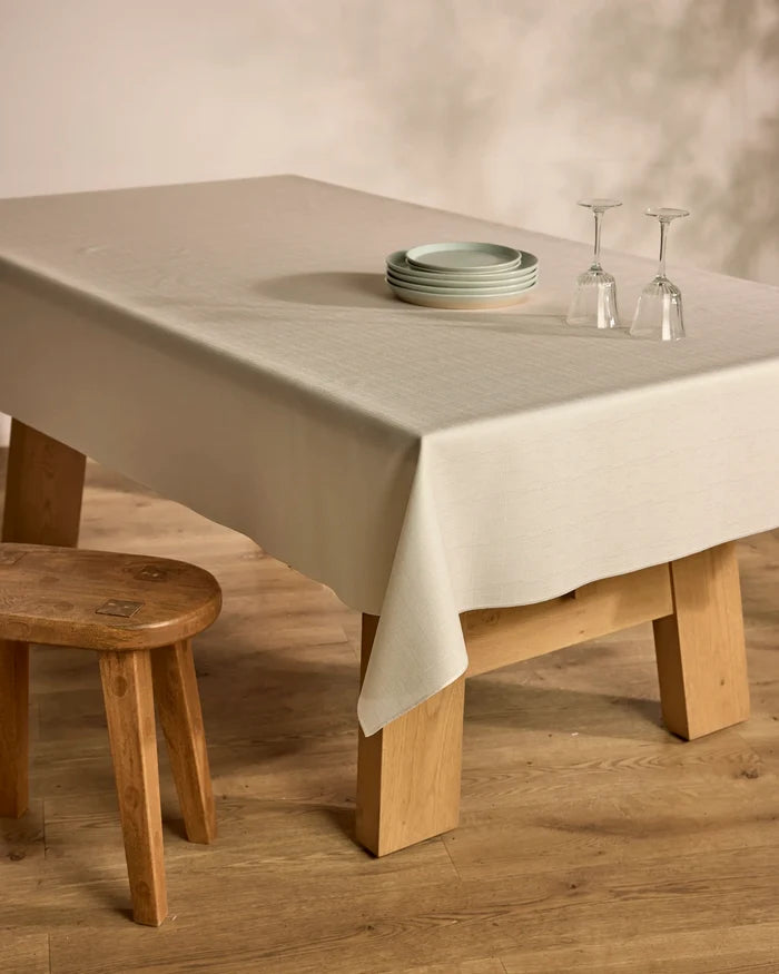 The Solid wooden table by Geometry, topped with a lightweight cloth, displays four stacked plates and two upside-down wine glasses. A matching wooden stool sits nearby on a wood floor against a neutral background.