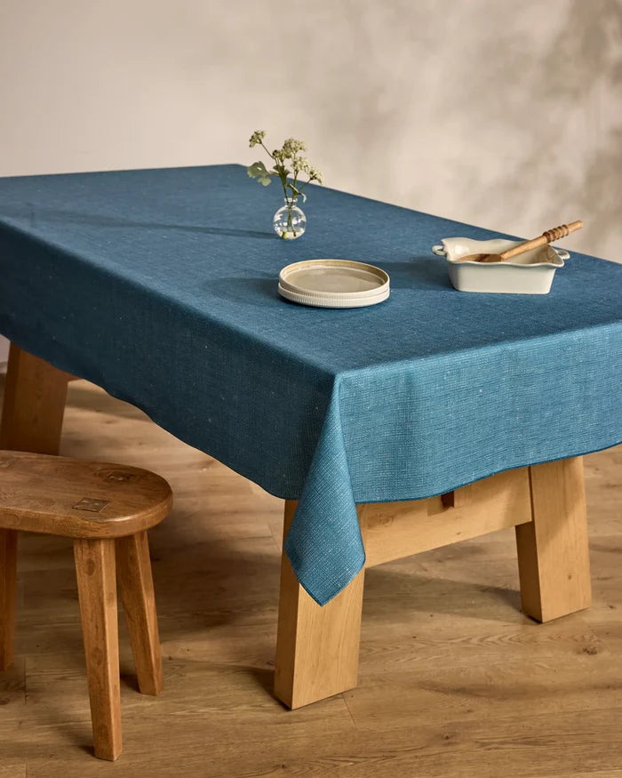A Geometry Solid wooden table with a lightweight blue tablecloth features a small round vase with white flowers, stacked white plates, and a white dish with a wooden utensil. A matching stool sits beside it on the wooden floor.