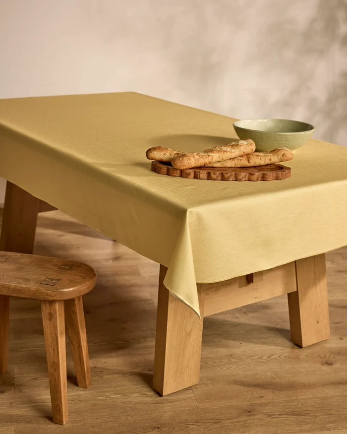 A Solid wooden table by Geometry, draped in a pale yellow cloth, features a tray of baguettes and a green bowl. Close by, a small wooden stool sits on the wood floor with a neutral wall as the backdrop.
