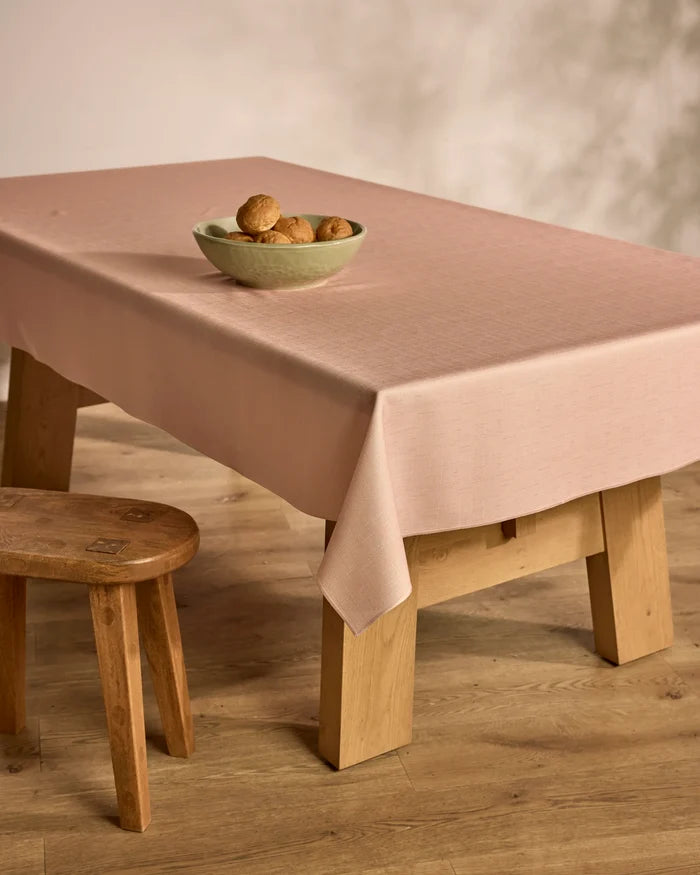 A Geometry Solid wooden table topped with a spill-resistant pale pink cloth holds a green bowl of brown rolls. A matching wooden stool sits beside it, all set on wood flooring against a light-colored wall.