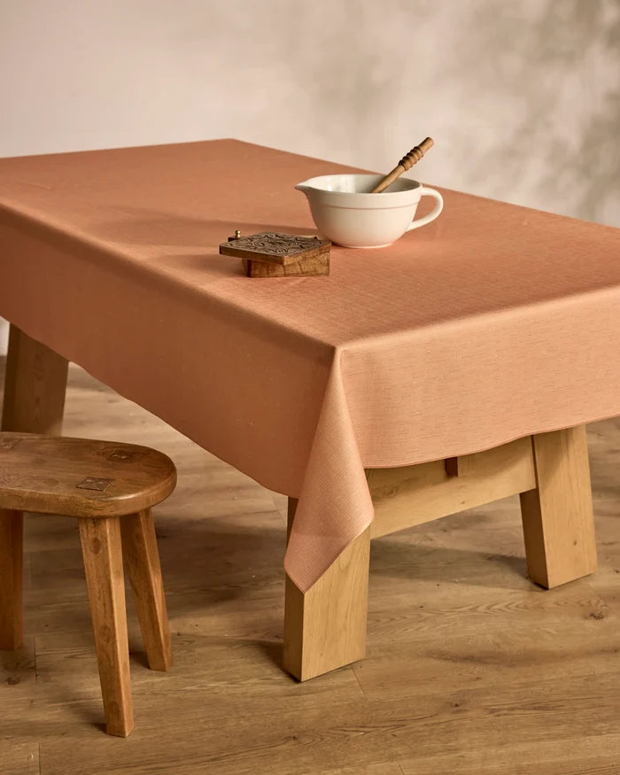 A Geometry Solid table is draped with a modern peach tablecloth, topped with a white mixing bowl, wooden spoon, and small brown book. A wooden stool sits close by on the wooden floor.