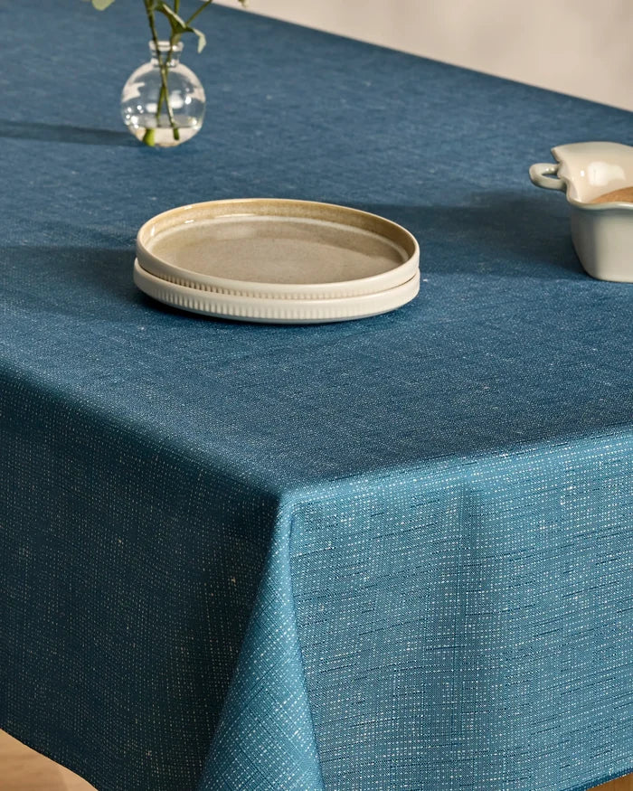 A Geometry Solid blue spill-resistant tablecloth covers a table set with a beige plate and small cream jug. A glass vase with a single flower is partially visible in the background.