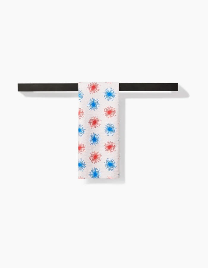 The Kayla Ann Sparkler Splash bar towel, crafted from Geoweave fabric with red and blue starburst patterns, hangs neatly over a black rod against a plain white background.