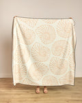 A person holds up the Spira beach blanket by Geometry, featuring a quick-drying cream fabric with a light tan nautilus shell pattern; only their hands and sandal-clad feet show against a plain wall and wooden floor.
