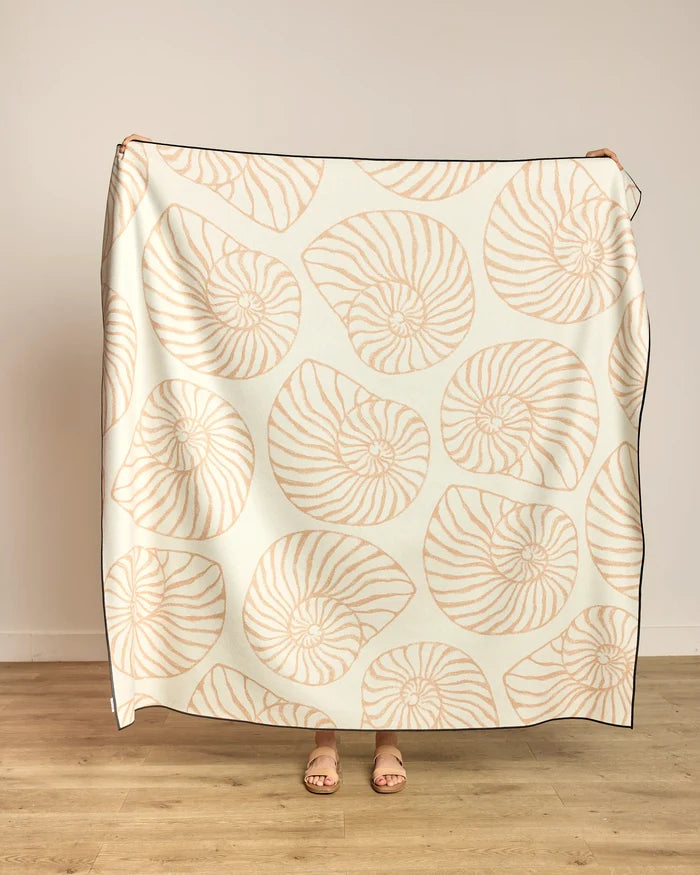 A person holds up the Spira beach blanket by Geometry, featuring a quick-drying cream fabric with a light tan nautilus shell pattern; only their hands and sandal-clad feet show against a plain wall and wooden floor.