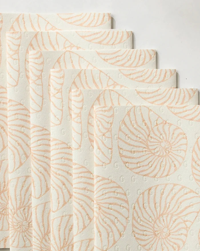 Spira by Geometry features five overlapping eco-friendly paper napkins with a pale cream background, a light peach seashell pattern, and a subtle embossed “G” design throughout the surface.