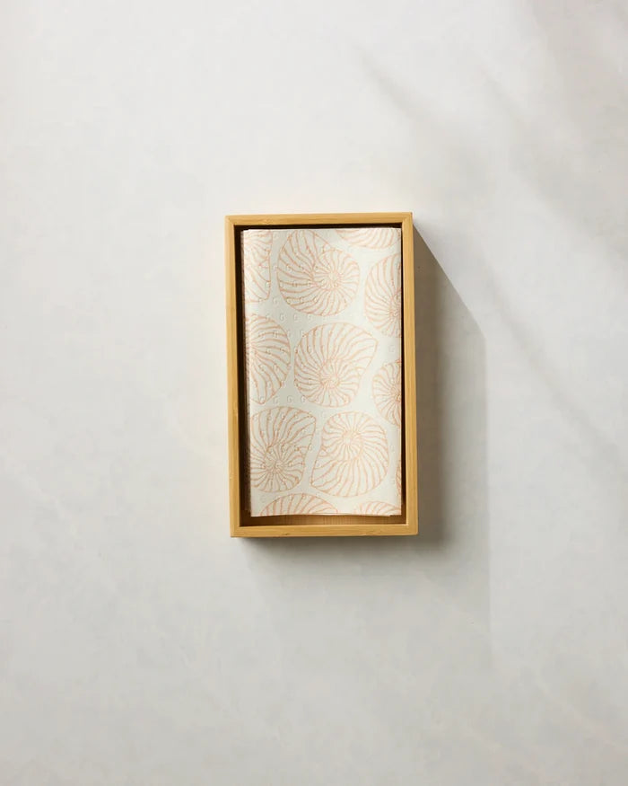 The Spira reusable paper towel by Geometry, featuring a light beige leaf pattern, is neatly folded inside a rectangular wooden tray on a light-colored textured surface.