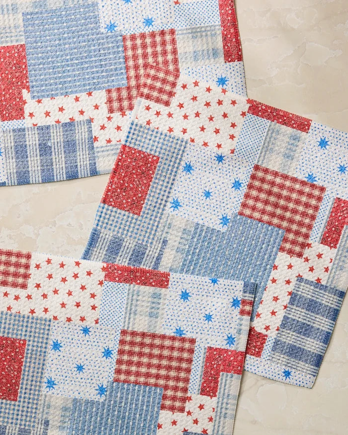 Three Stars and Stitches placemats by Geometry, featuring a red, white, and blue patchwork of checks, stripes, and stars, are displayed on a light beige marble-like surface—ideal to pair with your favorite dishcloth set.
