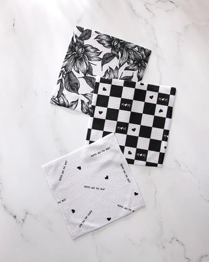 Three super-absorbent GEOMETRY "The Best" dishtowels rest on white marble: one has a black floral design, another a buffalo check with hearts, and the third features scattered text and tiny hearts—all made from recycled materials.