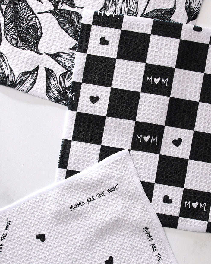 The GEOMETRY "The Best" set features three super-absorbent dish towels: one with black leaves, one black-and-white checkerboard with hearts and "MOM," and one scattered with "MOMS ARE THE BEST"—perfect for any kitchen.
