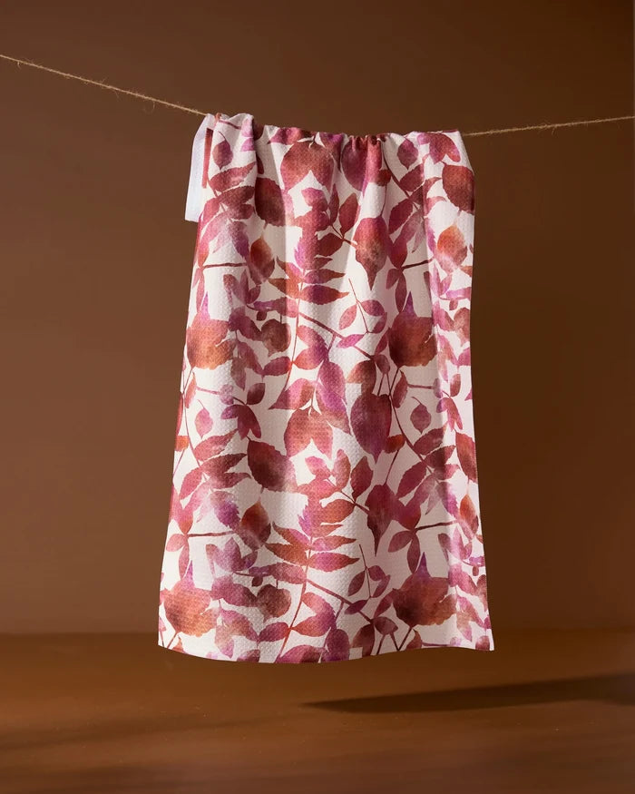 Geometry’s Watercolor Dreaming tea towel, featuring a pink and brown leaf pattern and made from recycled materials, hangs on a string against a brown background.