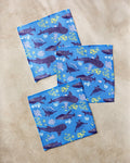 Three blue Whale Shark dishtowels by Elisabeth Wing, featuring whales, sharks, dolphins, and coral patterns, are displayed on a beige surface.