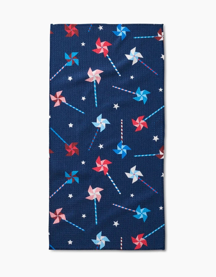 The Whirlwind Pins bar towel by Rebecca Bobko is navy blue, made from recycled materials, and features a festive pattern of red, white, and blue pinwheels with small white stars.
