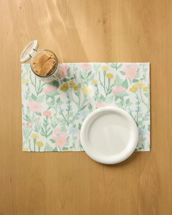 The "Wildflowers in Bloom" placemat by Jasmine Blooms Designs sits on a wooden table, holding a white bowl and a glass jar with a hinged lid filled with cookies.