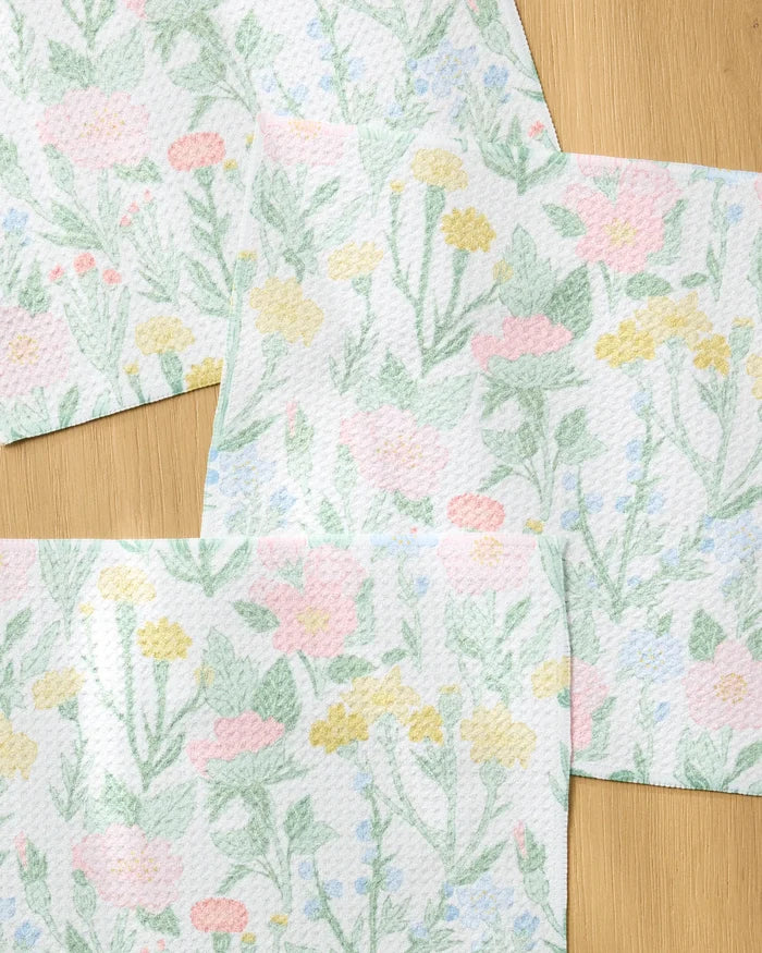 Wildflowers in Bloom paper towels by Jasmine Blooms Designs, made from recycled materials, showcase pastel pink, yellow, and green flowers and leaves overlapping on a light wood surface.