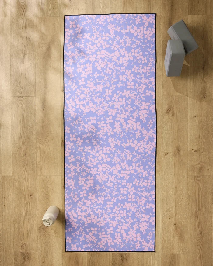 The Sareka Unique "You Give Me Butterflies" purple yoga mat with a pink abstract pattern is spread on a wooden floor, topped with a non-slip towel. A white water bottle sits on the left, and two gray yoga blocks are stacked on the upper right.